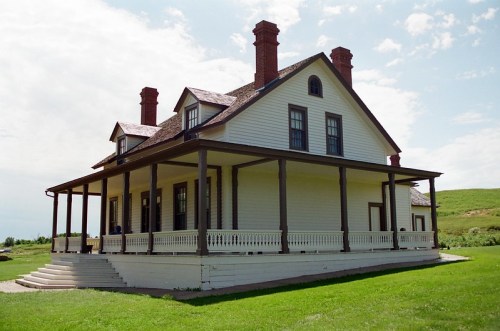 A large, two-story white house with a porch, featuring multiple chimneys and a green lawn under a cloudy sky.