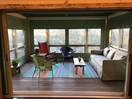 Cozy screened-in porch with a sofa, chairs, and a colorful rug, surrounded by large windows and plants.
