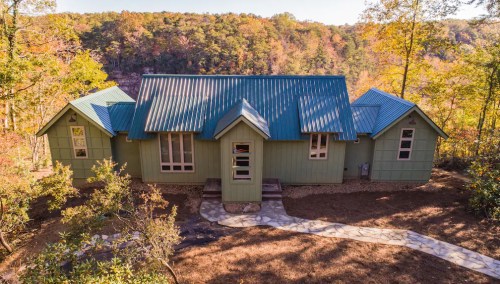A green house with a metal roof, surrounded by trees and autumn foliage, set against a hilly landscape.