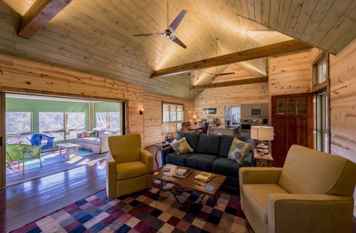 Cozy living room with wooden walls, a sofa, armchairs, and large windows showcasing a scenic view.