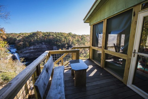 A wooden deck with a bench overlooks a scenic view of a waterfall and lush greenery on a clear day.