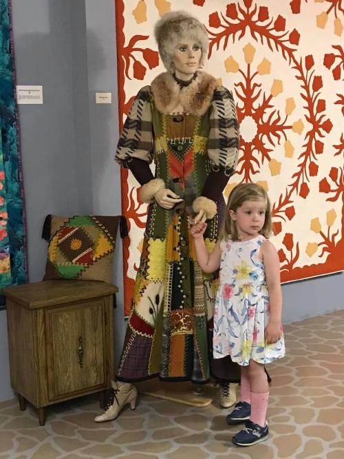 A child in a floral dress stands next to a mannequin dressed in a colorful patchwork outfit, with quilts in the background.