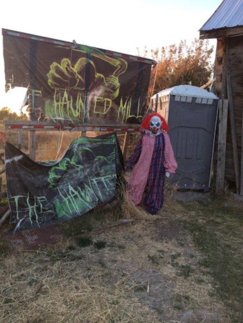 A creepy clown stands next to a "Haunted Mill" sign, with a portable restroom in the background and dry grass around.
