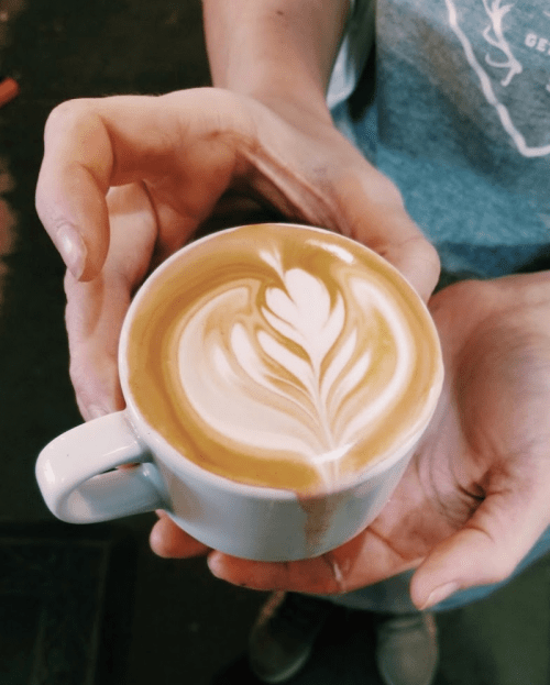 A person holding a white cup of latte art featuring a leaf design on top of creamy coffee.