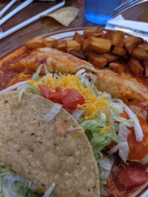 A plate of food featuring a taco with lettuce, tomato, and cheese, alongside enchiladas and seasoned potato wedges.