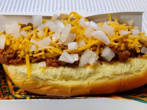 A close-up of a hot dog topped with chili, shredded cheese, and diced onions in a takeout container.