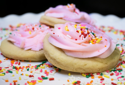 Three frosted cookies with pink icing shaped like roses, surrounded by colorful sprinkles on a white plate.