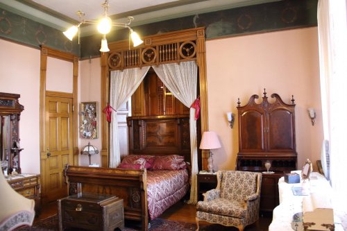 A vintage bedroom featuring a wooden bed with drapes, a patterned armchair, and antique furniture against pastel walls.