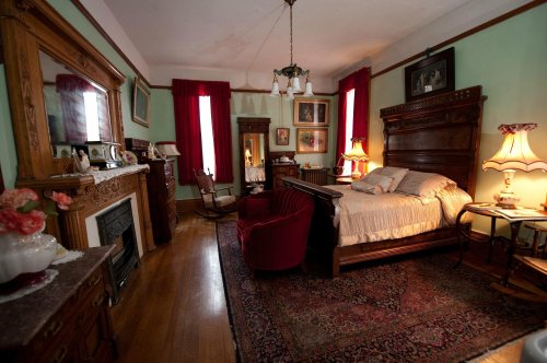 A vintage bedroom with a wooden bed, red curtains, ornate furniture, and a decorative mirror. Warm lighting creates a cozy atmosphere.