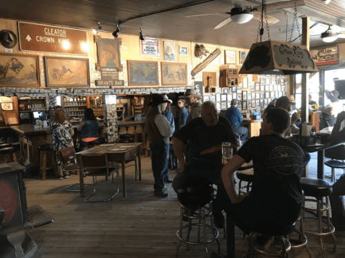 A rustic bar interior with patrons socializing, vintage decor, and cowboy-themed artwork on the walls.