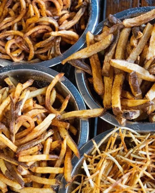 A variety of crispy fries in metal bowls, including curly fries and straight-cut fries, seasoned and golden brown.