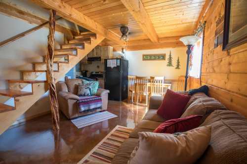 Cozy cabin interior featuring a living area, dining space, wooden stairs, and a kitchen in warm, rustic tones.