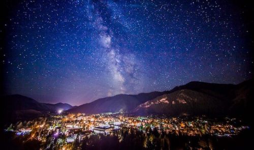 A starry night sky over a small town nestled in mountains, illuminated by city lights below.