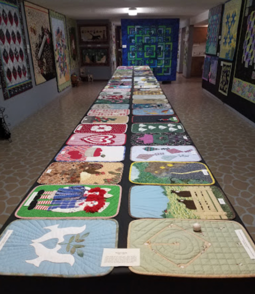 A long table displays various colorful quilts in a gallery setting, showcasing intricate designs and patterns.