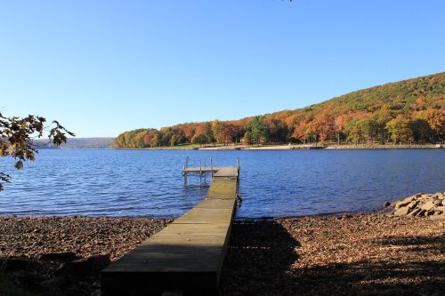 A wooden dock extends into a calm lake, surrounded by colorful autumn foliage on the hills in the background.