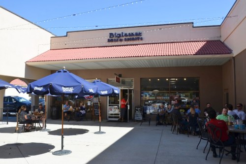 A café with outdoor seating, featuring blue umbrellas and patrons enjoying their time on a sunny day.