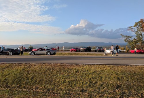 A scenic overlook with several cars parked along the road, and people enjoying the view of mountains and sky.