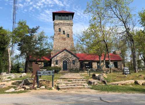 A stone building with a red roof and tower, surrounded by trees and signs, on a clear day.