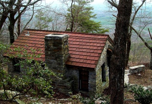 A small stone cabin with a red roof nestled among trees, overlooking a misty valley.