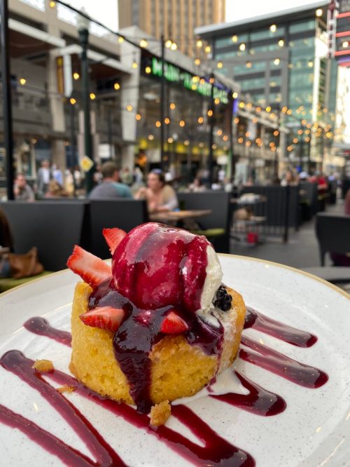 A slice of cake topped with strawberries, ice cream, and berry sauce, set against a lively outdoor dining scene.