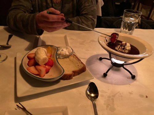 A hand holds a skewer over a chocolate fondue, with fruit, whipped cream, and bread on the plate beside it.