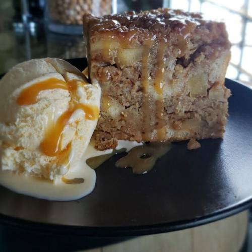 A slice of apple pie topped with caramel sauce, served alongside a scoop of vanilla ice cream on a black plate.