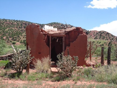 A weathered, crumbling adobe structure surrounded by desert vegetation and hills under a blue sky.