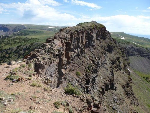 A rocky cliff edge overlooking a lush green landscape under a blue sky with scattered clouds.