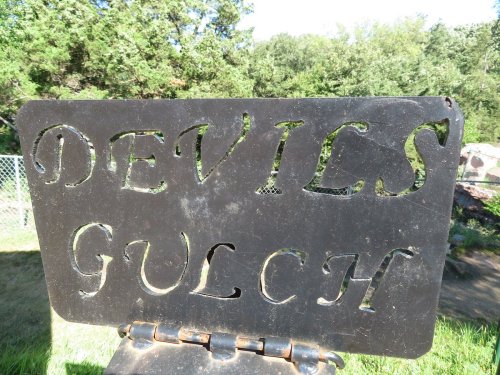 Sign reading "DEVILS GULCH" made of metal, set against a backdrop of trees and greenery.