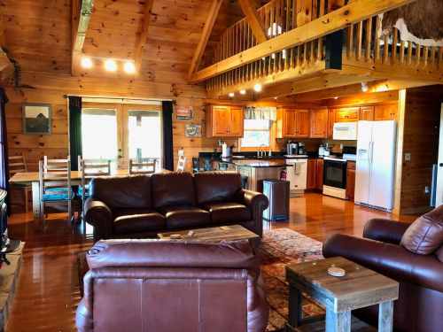Cozy cabin interior with wooden walls, leather sofas, a kitchen, and a loft area, featuring warm lighting and rustic decor.