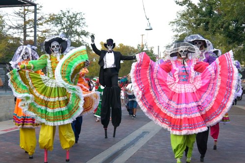 Colorful costumed performers with skull makeup parade down a street, showcasing vibrant dresses and festive attire.