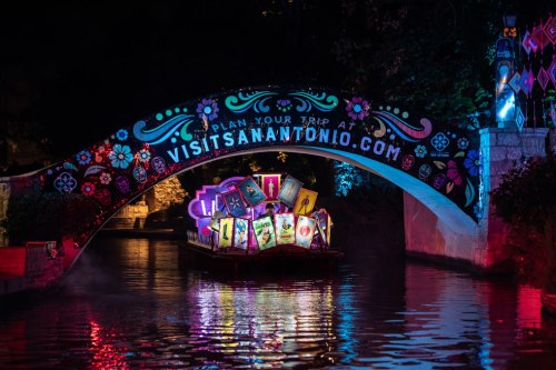 A colorful illuminated bridge over a river, with a boat displaying vibrant artwork and a website link for San Antonio.