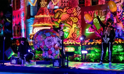 Colorful Day of the Dead celebration with skeleton performers, vibrant decorations, and a large decorated skull.