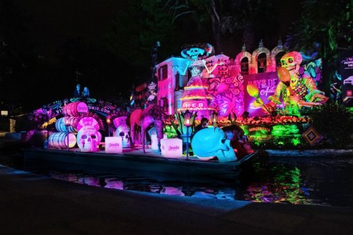 A colorful boat with large skull decorations floats on a river, surrounded by vibrant, illuminated murals at night.