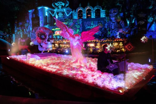 A vibrant float decorated with flowers, a skull, and an angel, illuminated in colorful lights for a Day of the Dead celebration.