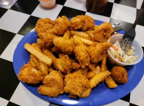A blue plate filled with golden fried shrimp, crinkle-cut fries, coleslaw, and a hushpuppy on a checkered table.