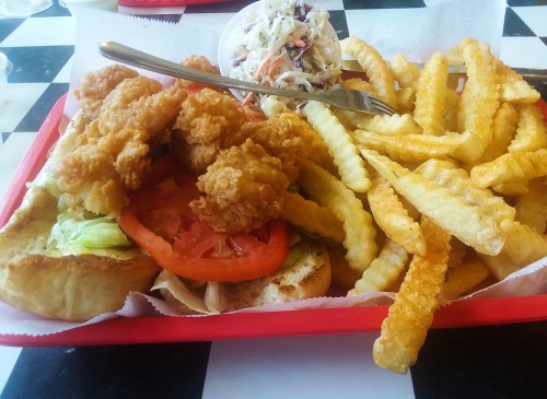 A red tray with a sandwich topped with fried shrimp, lettuce, and tomato, alongside crinkle-cut fries and coleslaw.