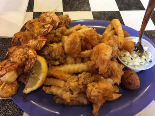 A plate of fried shrimp, crinkle-cut fries, hushpuppies, and coleslaw on a checkered tablecloth.