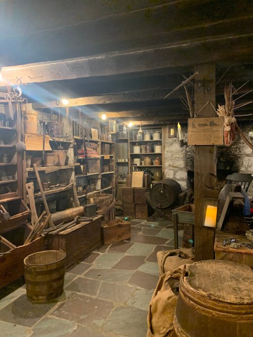 A rustic workshop filled with shelves of tools, barrels, and various containers, illuminated by warm overhead lights.