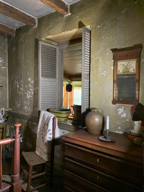 A rustic room with peeling green walls, a window with shutters, a wooden chair, and a candle on a dresser.