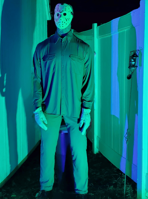 A tall figure in a hockey mask and gloves stands in a dimly lit outdoor area, casting a long shadow.