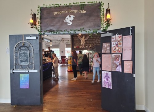 Entrance to Dragon's Forge Cafe, featuring a sign, decorative plants, and patrons inside a cozy, inviting space.