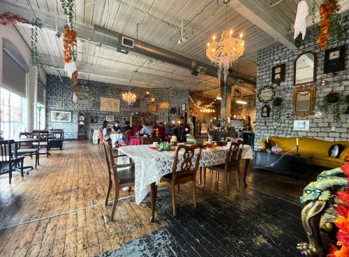 A cozy restaurant interior with wooden floors, decorative tables, and vintage decor, featuring chandeliers and plants.