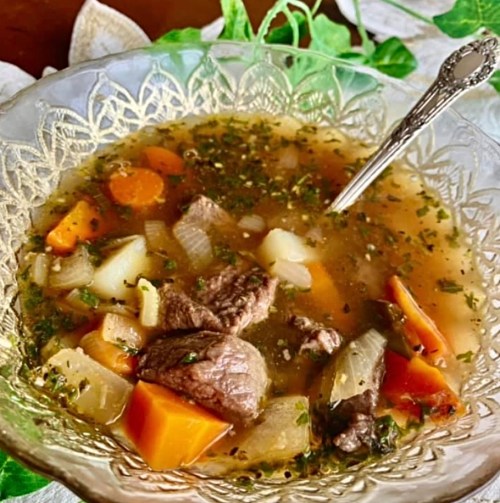A bowl of hearty beef soup with vegetables like carrots, potatoes, and herbs, served with a silver spoon.