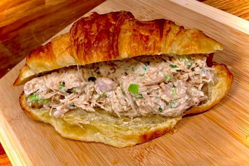 A flaky croissant filled with creamy chicken salad, garnished with green onions, on a wooden cutting board.