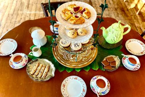 A tiered stand with assorted pastries, tea cups, and a teapot on a wooden table, surrounded by plates and a green leaf mat.
