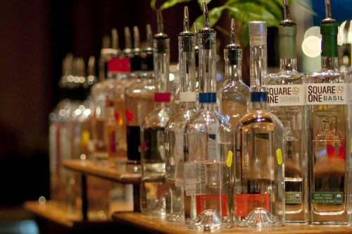 A row of clear liquor bottles on a wooden bar, with various labels and colors, illuminated by soft lighting.