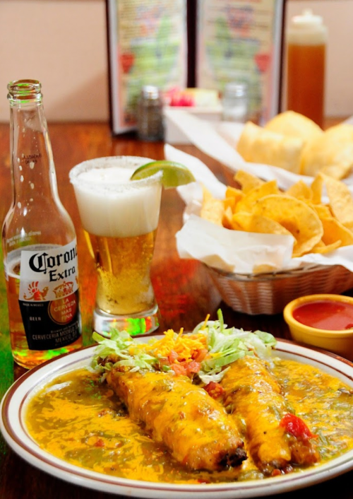 A plate of enchiladas topped with green sauce, alongside chips, a beer, and a glass of beer with lime.