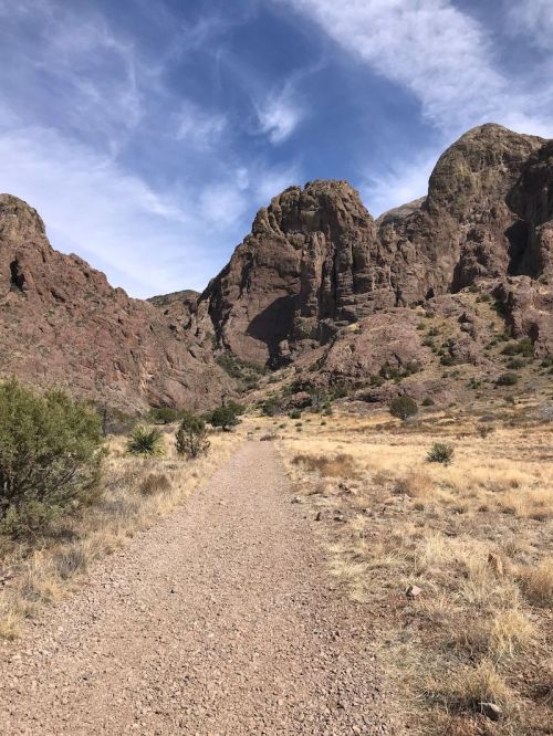 A rocky path leads through a dry landscape with towering mountains under a blue sky with scattered clouds.