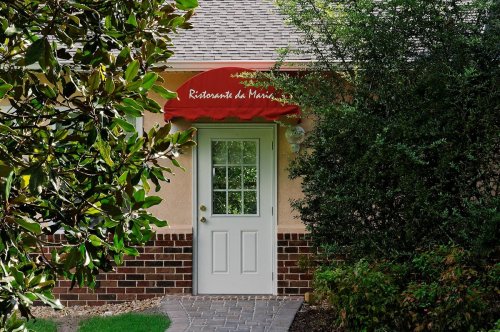 A restaurant entrance with a red awning, surrounded by greenery and a brick pathway.